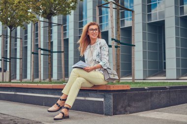 Bir gökdelen karşı parkta bir bankta oturmuş bir kitapla eğitim modern giysiler, çekici sarışın kadın.