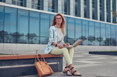 Şık moda blogger bir gökdelen karşı bir bankta oturmuş, dizüstü bilgisayar üzerinde çalışan açık rahatlatıcı.