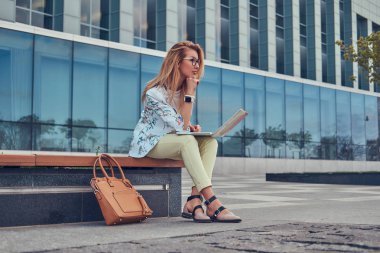 Şık moda blogger bir gökdelen karşı bir bankta oturmuş, dizüstü bilgisayar üzerinde çalışan açık rahatlatıcı.