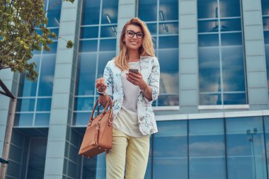 Güzel moda kadın blogger şık giysiler ve gözlük bir çanta ile gülümseyerek, bir gökdelen karşı duran bir akıllı telefon tutar.
