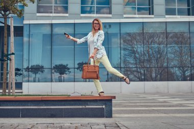 Komik güzel moda kadın blogger şık giysiler ve gözlük bir çanta ile tutan bir gökdelen karşı duran bir akıllı telefon.