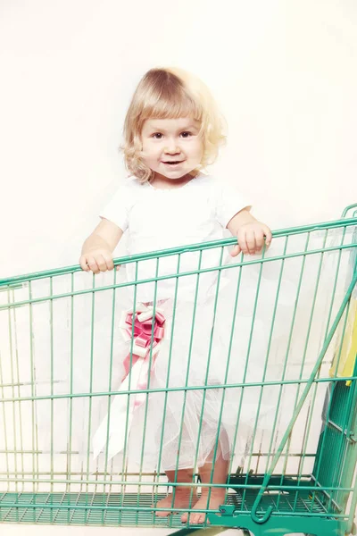 Retrato de uma menina bonita feliz em um vestido branco sentado em um carrinho com compras no fundo branco . — Fotografia de Stock