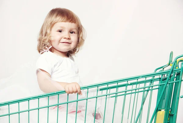 Retrato de uma menina bonita feliz em um vestido branco sentado em um carrinho com compras no fundo branco . — Fotografia de Stock