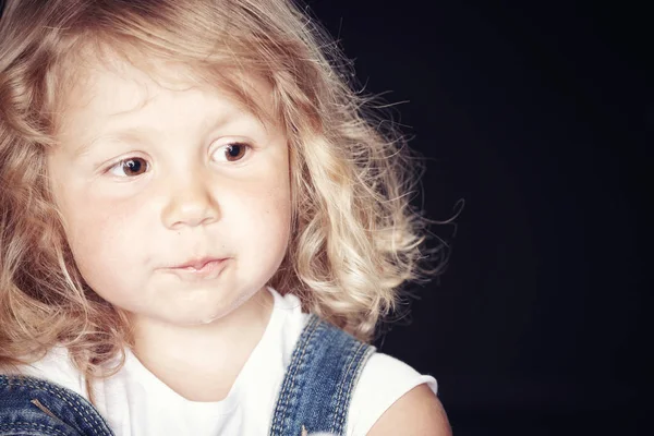 Портрет задумчивой милой маленькой девочки в джинсовом комбинезоне, сидящей в студии на черном фоне . — стоковое фото