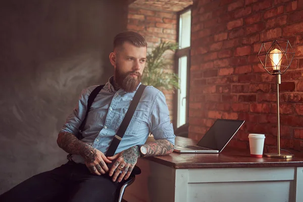 Όμορφος τατουάζ hipster σε ένα πουκάμισο και τιράντες που κάθεται στο γραφείο με έναν υπολογιστή, κοιτάζοντας έξω από το παράθυρο σε ένα γραφείο με εσωτερικό πατάρι. — Φωτογραφία Αρχείου