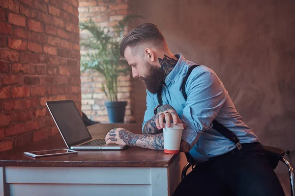 Hezký Tetovaný bokové v košili a podvazky, sedí u stolu, pracují na notebooku, drží s sebou kávu v kanceláři s interiér podkroví. — Stock fotografie