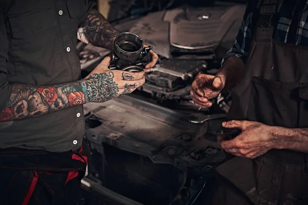 Deux mécaniciens examinent un détail du moteur alors qu'ils se tiennent près d'une voiture cassée dans un garage . — Photo