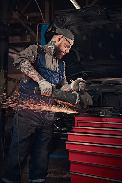 Mécanicien automobile brutal dans un uniforme et lunettes de sécurité travaillant avec une meuleuse d'angle tout en se tenant contre une voiture cassée dans le garage de réparation . — Photo