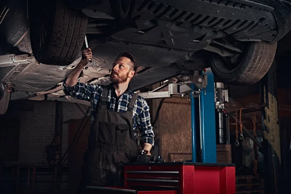 Brodaty auto mechanik w mundurze naprawy zawieszenia samochodów z kluczem dynamometrycznym, stojąc pod podnoszenia samochodu w garażu napraw. — Zdjęcie stockowe