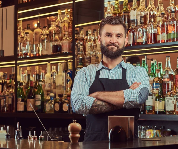 Stylish brutal barman in a shirt and apron standing with crossed arms at bar counter background.