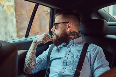Lüks bir araba bir arka koltukta oturan askısı ile bir gömlek dalgın eski moda dövmeli hipster adam uzağa arıyorsunuz.