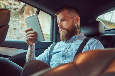 Eski moda dövmeli hipster erkekte bir arka koltukta bir lüks arabada otururken bir tablet kullanarak bir gömlek askısı,.