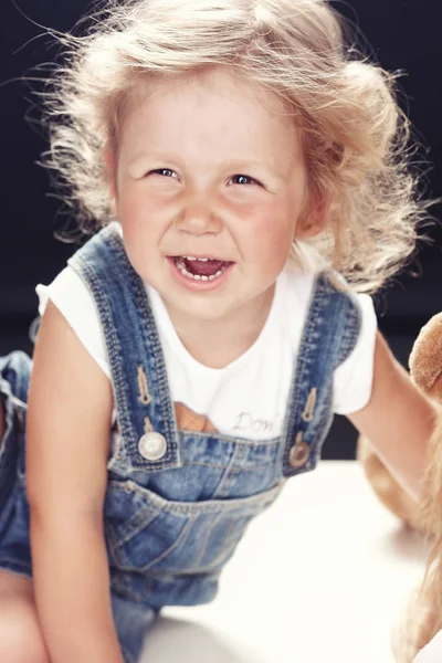 Porträt eines aufgebrachten kleinen Mädchens in Jeans-Overalls, das in einem Studio auf schwarzem Hintergrund sitzt. — Stockfoto