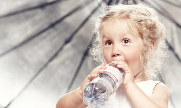 Portrét srandovní roztomilé malé holčičky v neformální oblečení, pít vodu, zatímco sedí v studio. — Stock fotografie