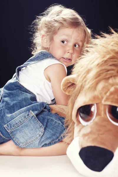 Portrait of a scared little girl in denim overalls, sitting in a studio on black background. — Stock Photo, Image