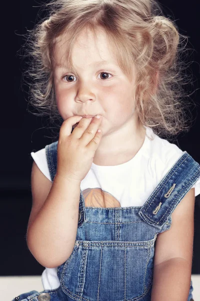 Portrait of a pensive cute little girl in denim overalls, sitting in a studio on black background. — Stock Photo, Image