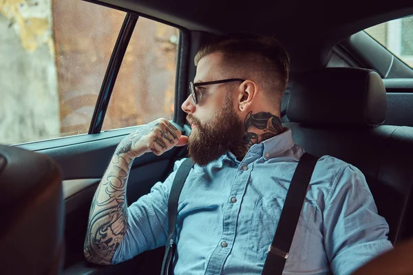 Pensive old-fashioned tattooed hipster guy in a shirt with suspenders sitting in a luxury car on a back seat, looking away.