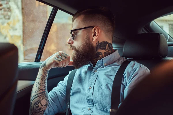 Pensive old-fashioned tattooed hipster guy in a shirt with suspenders sitting in a luxury car on a back seat, looking away.