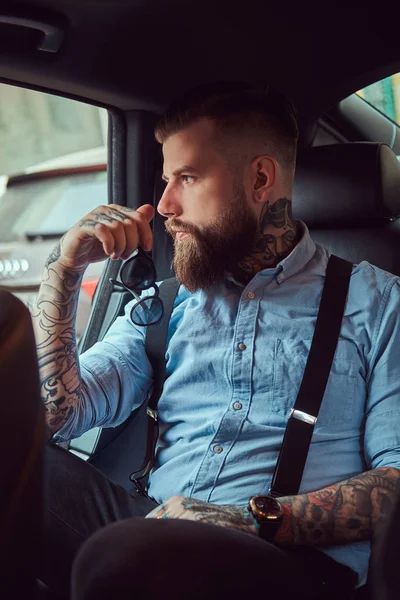 離れて見て高級車の後部座席に座っているサスペンダー付きシャツで物思いにふける昔ながらの刺青ヒップスター男 — ストック写真