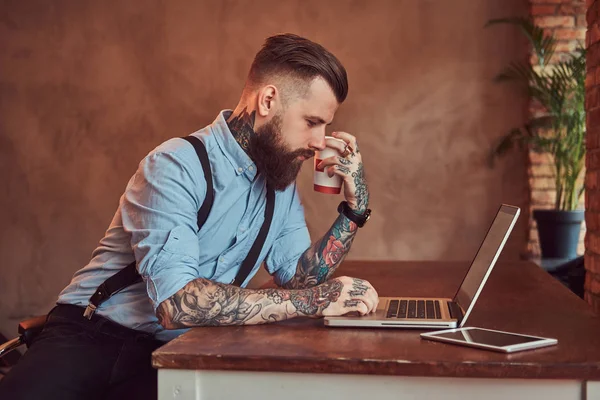 Bello hipster tatuato in camicia e bretelle seduto alla scrivania, che lavora su un computer portatile, tiene caffè da asporto in un ufficio con un interno soppalco . — Foto Stock