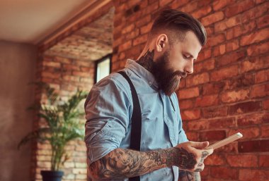 Çatı katı iç bir ofiste bir resepsiyon yakın dururken tablet kullanma bir gömlek ve pantolon askısı, bir güneş gözlüğü takan eski moda dövmeli hipster.