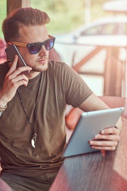 Yakışıklı şık hipster telefonda konuşurken yol kenarına yerleştirilen bir kafede bir masada oturur ve tablet tutar.