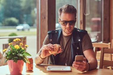 Bir hamburger yerken bir smartphone kullanarak bir şık saç ve sakal ile yakışıklı hipster yol kenarına yerleştirilen bir kafede yemek yemeye karar verdim bir masada oturur.