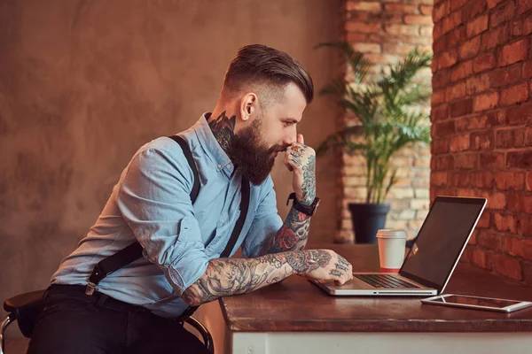 Bellissimo hipster tatuato in camicia e bretelle seduto alla scrivania, che lavora su un computer portatile, in un ufficio con interni a soppalco . — Foto Stock