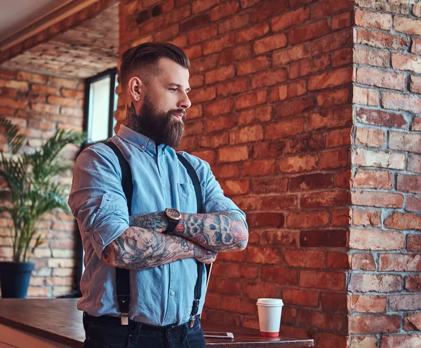 Old-fashioned tattooed hipster in a shirt and suspenders, standing near a desk with a laptop, looking out the window in an office with loft interior.