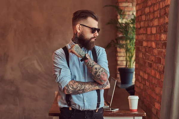 Hipster tatuado a la antigua con una camisa y tirantes, en gafas de sol, de pie con los brazos cruzados cerca de un escritorio con un ordenador portátil, mirando por la ventana en una oficina con un interior loft . —  Fotos de Stock