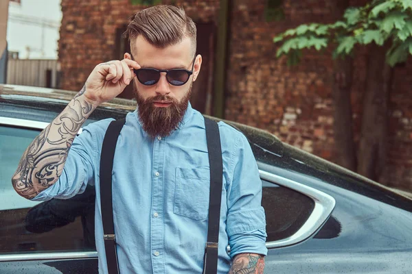 Old-fashioned tattooed hipster guy in a shirt with suspenders, standing near black car outdoors.