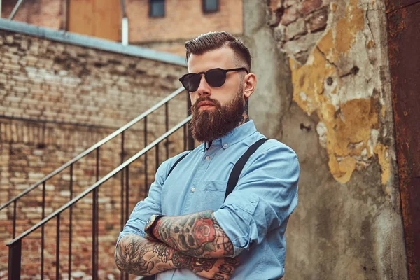 Élégant beau vieux tatoué hipster guy dans une chemise avec des bretelles, debout près d'un vieux bâtiment à l'extérieur . — Photo
