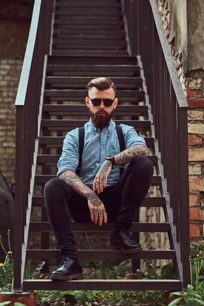 Elegante ragazzo hipster tatuato vecchio stile con una camicia con bretelle, seduto sulle scale vicino al vecchio edificio all'aperto . — Foto Stock