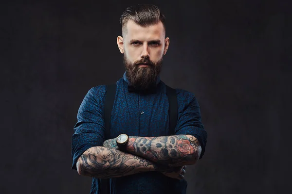 Portret van een knappe ouderwetse hipster in een blauw shirt en jarretels, permanent met gekruiste armen in een studio. — Stockfoto