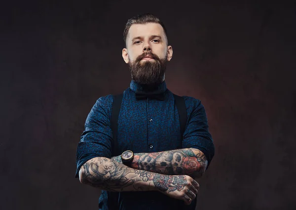 Retrato de un guapo hipster a la antigua con una camisa azul y tirantes, de pie con los brazos cruzados en un estudio . — Foto de Stock