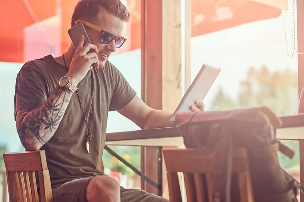 Bonito hipster elegante senta-se a uma mesa em um café à beira da estrada, falando ao telefone assistindo a um tablet . — Fotografia de Stock