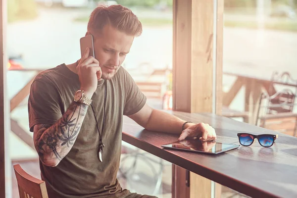 Όμορφος μοντέρνος hipster κάθεται σε ένα τραπέζι σε ένα καφέ άκρη του δρόμου, μιλώντας σε ένα smartphone. — Φωτογραφία Αρχείου