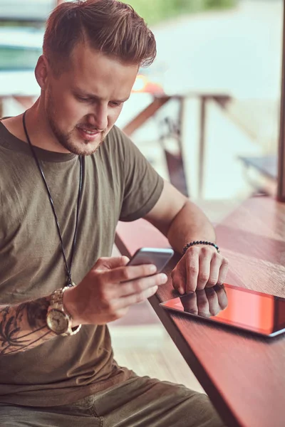 Όμορφος μοντέρνος hipster κάθεται σε ένα τραπέζι σε μια άκρη του δρόμου καφέ, διαβάζοντας το μήνυμα στο smartphone. — Φωτογραφία Αρχείου