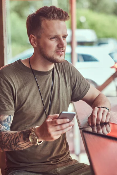 Knappe stijlvolle hipster zit aan een tafel in een café langs de weg, het bezit van een smartphone. — Stockfoto