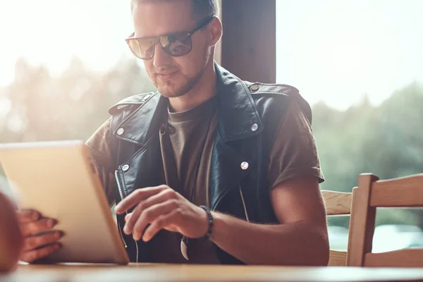 Knappe hipster met een stijlvol kapsel en baard zit aan een tafel in een café langs de weg, ziet er ongeveer in de tablet-pc. — Stockfoto