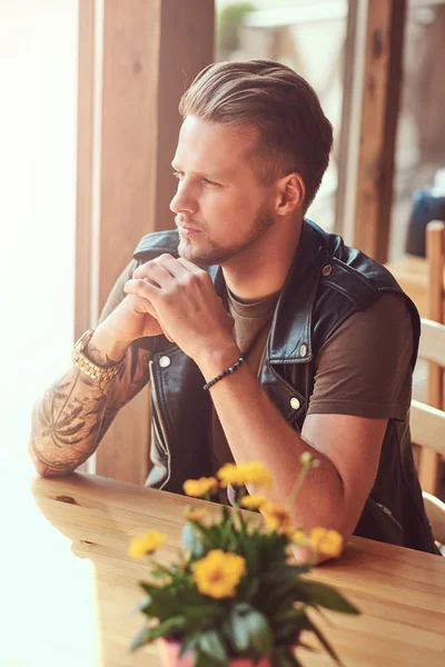 Hipster con un corte de pelo elegante y barba se sienta en una mesa en un café al lado de la carretera, mira a la calle . —  Fotos de Stock