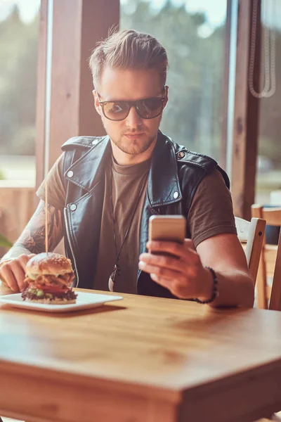 Hipster με μοντέρνο κούρεμα και γενειάδα που κάθεται σε ένα τραπέζι, χρησιμοποιώντας ένα smartphone κατά τη διάρκεια γεύματος σε οδική καφέ. — Φωτογραφία Αρχείου