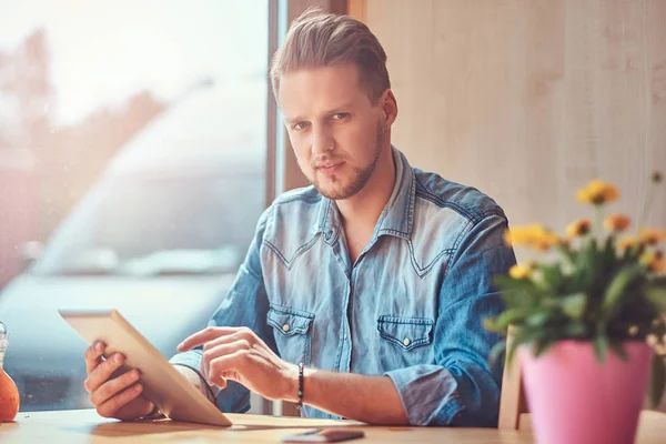 Hipster guy with a stylish haircut and beard sits at a table in a roadside cafe, holds a tablet computer. — Stock Photo, Image