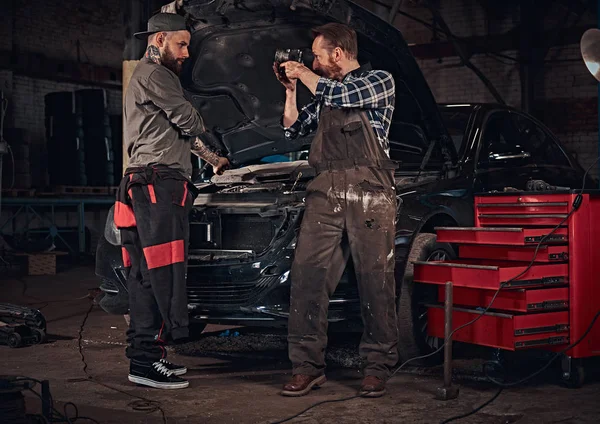 Deux mécaniciens barbus en uniforme, réparer une voiture cassée dans le garage . — Photo