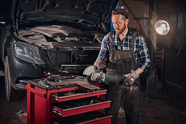 Mécanicien automobile brutal dans un uniforme et lunettes de sécurité travaillant avec une meuleuse d'angle tout en se tenant contre une voiture cassée dans le garage de réparation . — Photo