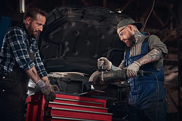 Deux mécaniciens barbus dans un uniforme et des lunettes de sécurité travaillant avec une meuleuse d'angle tout en se tenant contre une voiture cassée dans le garage de réparation . — Photo