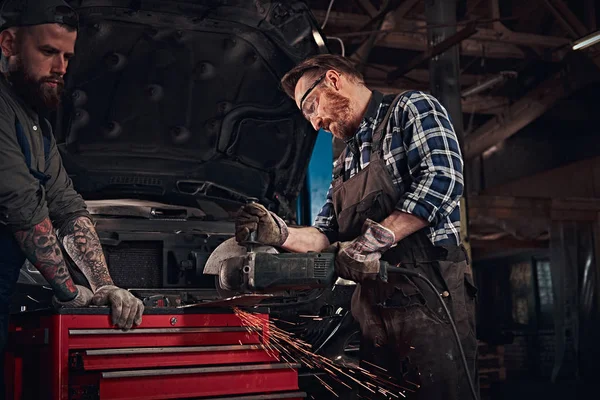 Deux mécaniciens barbus dans un uniforme et des lunettes de sécurité travaillant avec une meuleuse d'angle tout en se tenant contre une voiture cassée dans le garage de réparation . — Photo