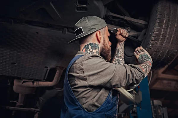 Bearded auto mechanic in a uniform repair the cars suspension with a wrench while standing under lifting car in repair garage.