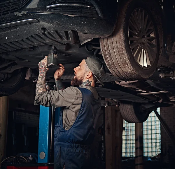 Auto mechanik w jednolity, naprawić samochód śrubokrętem stojąc pod podnoszenia samochodu w garażu napraw. — Zdjęcie stockowe