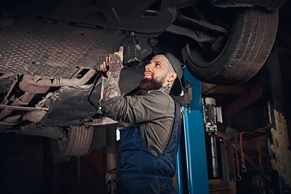 Auto mechanic in a uniform, repair the car with a screwdriver while standing under lifting car in the repair garage.